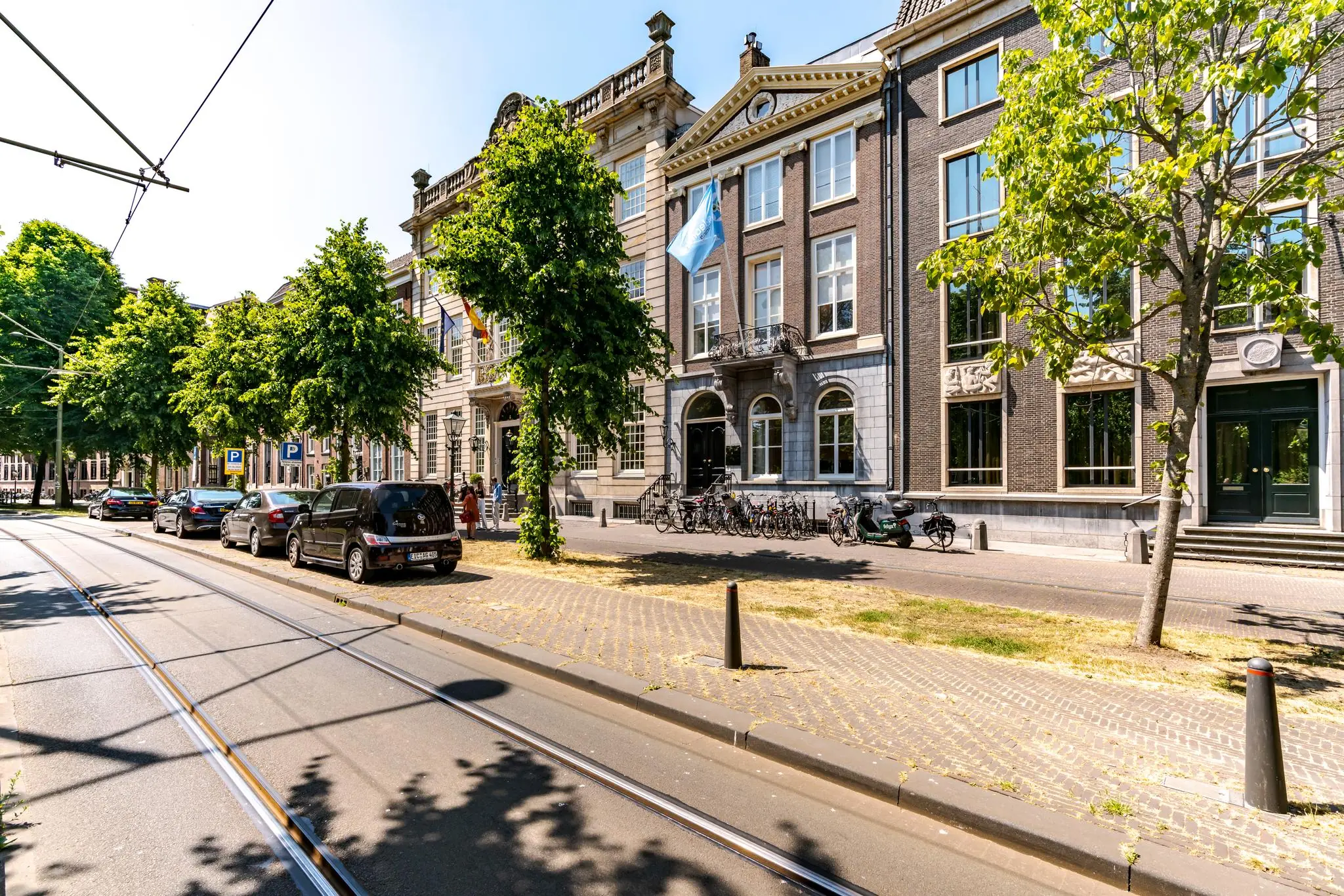 Zonnige straat met historische gebouwen aan de Lange Vijverberg in Den Haag, met geparkeerde auto's, fietsen en tramlijnen op de voorgrond.